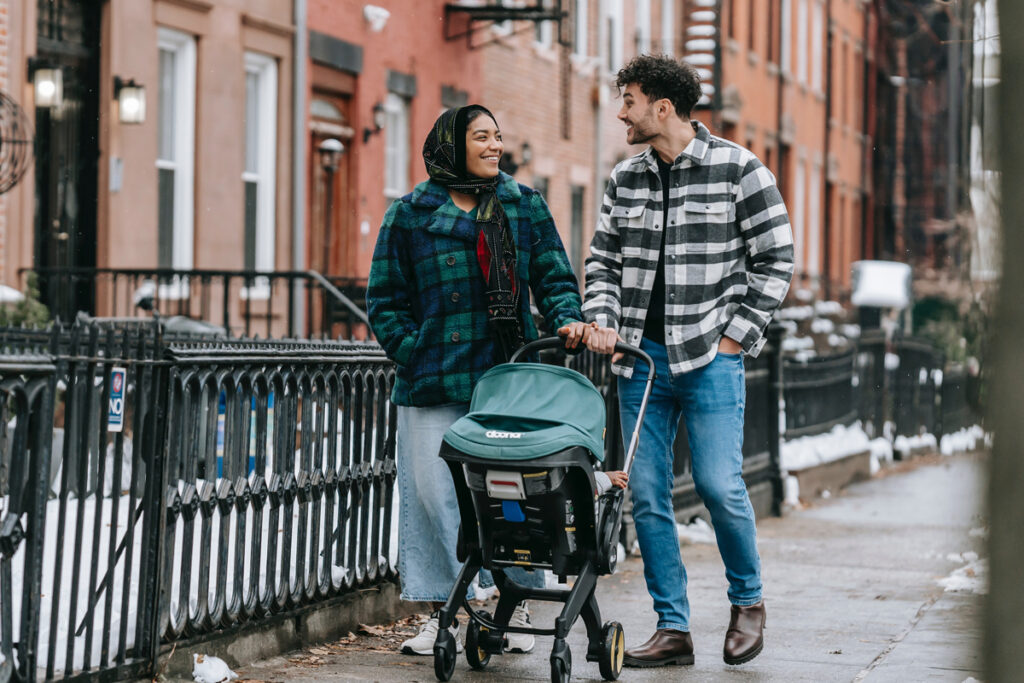 Une mère et un père promènent leur bébé en poussette dans la ville. Ils sourient et semblent heureux. / article bébé seconde main