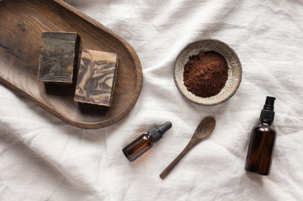 Un savon solide est placé sur un drap blanc avec à côté une coupelle avec de la poudre marron, une cuillère et des produits de beauté. / produits cosmétiques naturels