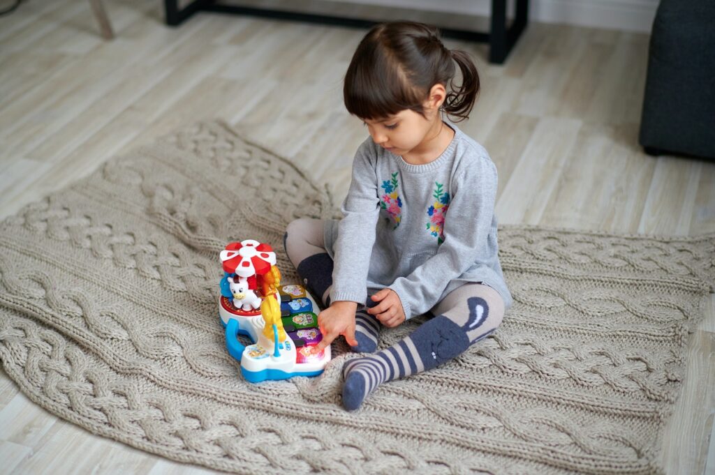 Une petite fille assise sur le sol avec une couverture, joue avec un piano musical en plastique. / marques de jouets écologiques
