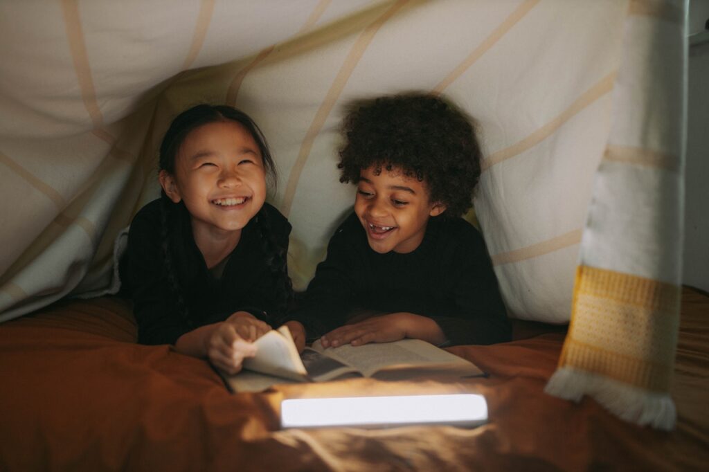 Une petite fille et un petit garçon sont souriants sous une tente, éclairée à la lampe de poche, ils regardent des livres. / recycler ses vieux livres